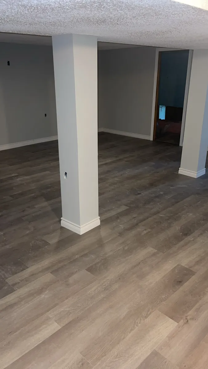 Grey plank flooring laid through a finished basement, with the support posts trimmed in new white baseboard
